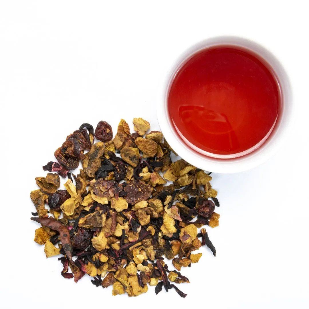 A cup of Cranberry Apple Tart Herbal Tea sits next to a loose pile of dried fruit and flower pieces on a white background.